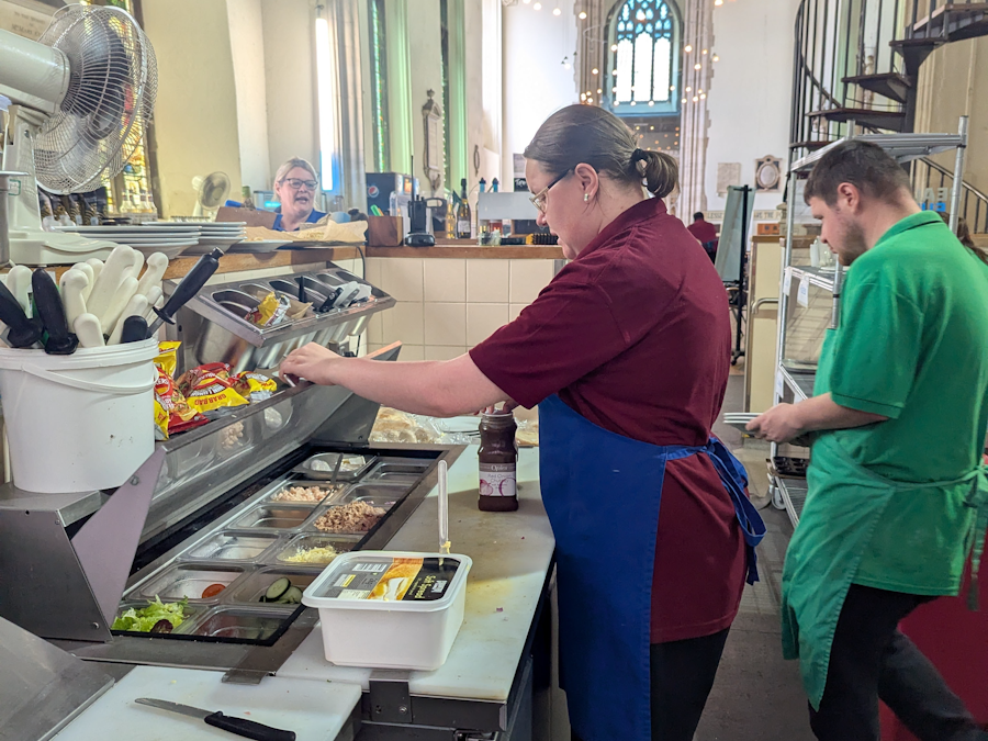 Café staff in aprons preparing food in the kitchen area at St Lawrence Café, with church arches visible in the background